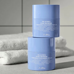 Two blue containers of Dr. Akehea Draelthea Pore Refresh Grinding Cleansing Balm on a light gray surface with a white towel in the background.