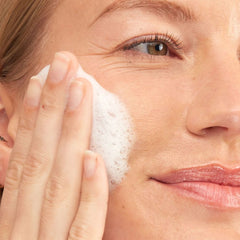 FANCL Aging Care Antioxidant Exfoliating Cleansing Cream[ Limited Edition] -THE COSMETIC STORE NZ-Close-up of a woman applying white foam cleanser to her cheek with her hand, showing her face and hand in warm, soft lighting.