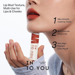 A close-up of a woman's lower face and hand holding a tube of lip mud. The product packaging is white with red stripes and vertical text reading 'IN TO YOU'.- INTO YOU SHERO SUPER MATTE LIP&CHEEK MUD 2G-EM08 BROWN-TAUPE/ BRICK RED NUDE-The Cosmetic Store