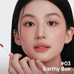 Close-up of a woman holding a lip product applicator near her lips, with text reading '#03 Earthy Box' in the lower right on a clean white background.