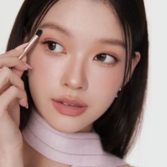 A close-up of a woman with dark hair and warm brown eyes, holding a makeup brush near her eye while wearing a pale pink top - Judydoll Mini Makeup Palette 02 Taro Box with mascara open showing mirror and mauve taro shades NZ