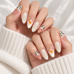 Close-up of hands with almond-shaped press-on nails featuring a mix of glossy white, sheer nude pink, black and white stripes, and small white daisy designs with yellow centers. The hands are resting on a white knit sweater.