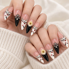 A close-up of manicured fingernails with a French tip design, featuring a gradient from pale pink to black, decorated with white floral motifs, small yellow flower centers, and silver rhinestone accents. The hands are resting on a soft cream-colored knit sweater.