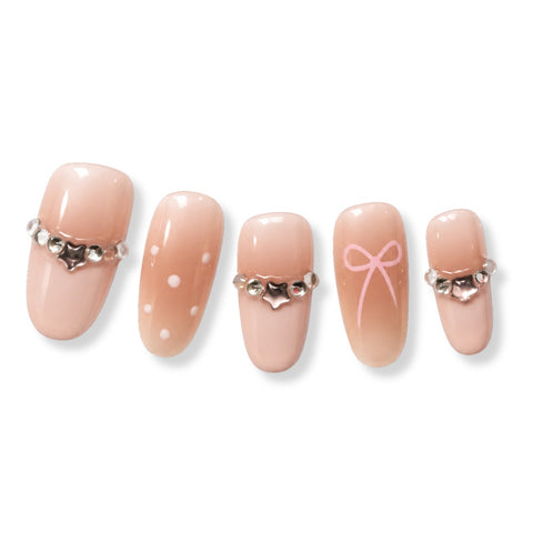 Five false press-on nails in a glossy pale peach pink shade, arranged horizontally on a white background. The nails feature different decorative designs: one with a silver rhinestone choker, one with white polka dots, one with a silver rhinestone bow, one with a pink bow print, and one with a silver rhinestone choker.