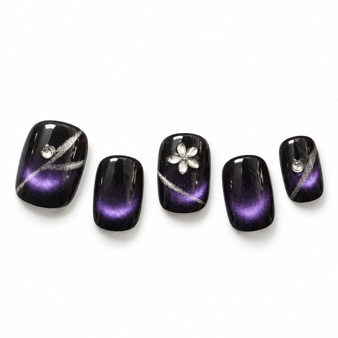 Five false press-on nails with a glossy black-to-purple cat-eye gradient design, decorated with silver glitter lines, a silver flower on one nail, and small clear rhinestones, displayed on a white background.