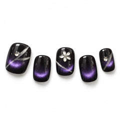 Five false press-on nails with a glossy black-to-purple cat-eye gradient design, decorated with silver glitter lines, a silver flower on one nail, and small clear rhinestones, displayed on a white background.