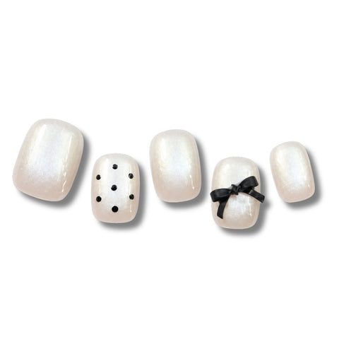 Five false press-on nails displayed on a white background, featuring a glossy pearlescent white finish with two accent nails decorated with black polka dots and a small black bow.