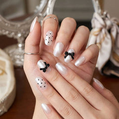 A close-up of manicured fingernails on a hand, featuring glossy white nails with black polka dots on two nails and two black bow designs on the other nails.