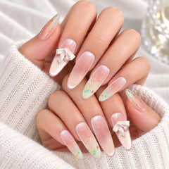 Press-on Nail |Fairycore Style |3D Ribbon Bows |Medium Almond Shap -A close-up of hands with long, glossy almond-shaped nails painted in a soft pink-to-white ombre style, with small white bow accents on two nails. The hands are posed on a beige fabric surface with decorative shell and feather props nearby.