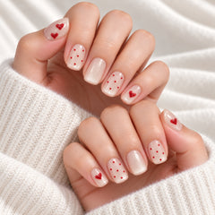 Press-on Nail |French Romantic Heart Print Style |Short Oval Shape- A close-up of hands showcasing a delicate nail art design with white and nude bases, red heart details, and red polka dots, styled with small glass vials and a pearl bracelet on a soft beige fabric background.