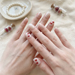 Press-on Nail |French Romantic Heart Print Style |Short Oval Shape- A close-up of hands showcasing a delicate nail art design with white and nude bases, red heart details, and red polka dots, styled with small glass vials and a pearl bracelet on a soft beige fabric background.