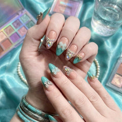 Press-on Nail | Y2K Turquoise French V-Tip Style | Almond Shape- A close-up of a hand with a decorative manicure featuring mint green and nude pink nails adorned with rhinestones, white bows, and butterfly embellishments, set against a turquoise satin background with eyeshadow palettes and bracelets.