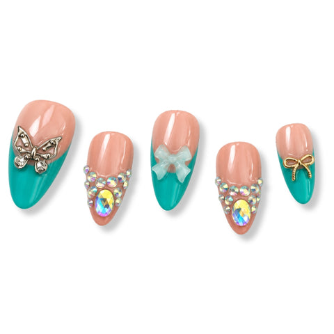 Five almond-shaped press-on nails with a peach and teal color-blocked design, decorated with rhinestones, a butterfly, a bow, and gold and iridescent accents, displayed on a white background.