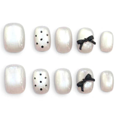 Ten false press-on nails arranged in two rows on a white background. The nails are glossy pearlescent white with a subtle iridescent sheen, with two nails decorated with small black polka dots and two decorated with black ribbon bows.