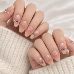 A close-up of manicured fingernails on hands, featuring a glossy nude-pink gradient base with small white dots, pink bow designs on two nails, and silver rhinestone accents. The hands are resting on a cream-colored knit sweater against a soft white fabric background.