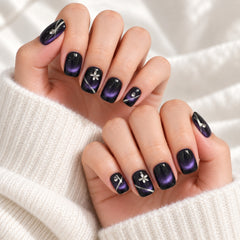 Close-up of hands with manicured nails painted in a glossy black-to-purple gradient with small silver floral and diagonal stripe accents, styled with a cream knit sweater.