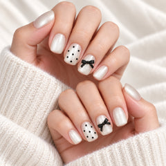Close-up of manicured hands wearing white press-on nails with some decorated in black polka dots and two black bows, styled with a cream knit sweater.