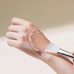 Hand applying a transparent gel-like substance to another hand using a brush on a neutral background