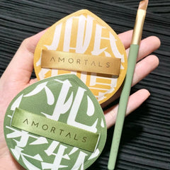 Two round makeup products with 'Amortals' branding held by a hand against a dark textured background.