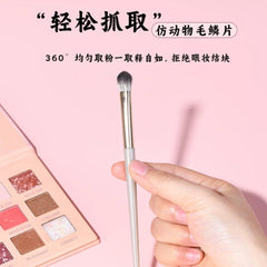 Makeup brush held by a hand with a pink makeup palette in the background