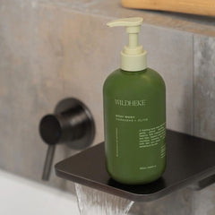 A bottle of Wildhike Body Wash in a dark green bottle with a cream-colored pump dispenser, resting on a black floating shelf in a modern bathroom setting with water running below.-THE COSMETIC STORE NZ.