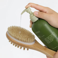 A hand holds a green pump bottle dispensing clear liquid onto a natural bamboo body brush with beige bristles, shown against a clean white background.-THE COSMETIC STORE NZ.