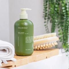 A green pump bottle of WILDHEKE body wash sits on a wooden tray beside a rolled white towel and a wooden scrub brush, with a blurred bathroom setting and hanging greenery in the background.- Close up of clear plant based Wildheke body wash lather with fresh native Harakeke flax leaves-THE COSMETIC STORE NZ.