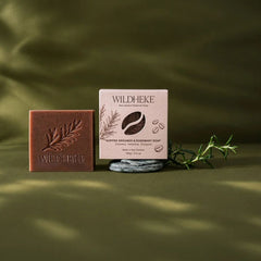 Wildheke natural exfoliating coffee soap bar 100g sitting on a rustic stone soap dish NZ- Wildheke Coffee Grounds & Rosemary soap set, featuring a rectangular brown bar of soap embossed with a leaf design and a matching light pink box, styled on a dark olive green background with a small green sprig.