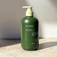 Wildheke Natural Conditioner Olive and Kawakawa 500ml pump bottle at The Cosmetic Store-A dark olive green pump bottle of Wildheke Conditioner with a cream-colored pump dispenser, standing on a stone surface against a neutral beige wall with soft sunlight and shadows.