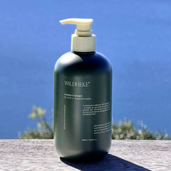 Wildheke Natural Conditioner Olive and Kawakawa 500ml pump bottle at The Cosmetic Store-A dark green pump bottle of WILDHEKE Conditioner Olive + Karakuma, set on a wooden surface with a blurred blue sky and foliage background.