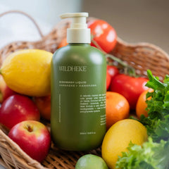 Wildheke Natural Dishwash Liquid Harakeke and Kawakawa 500ml pump bottle at The Cosmetic Store-A bottle of WILDHEKE Dishwash Liquid centered in a wicker basket, surrounded by fresh produce including lemons, apples, tomatoes, and greens.