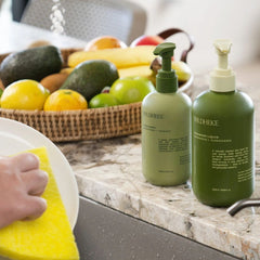 Two green Wildheke liquid hand wash bottles on a marble kitchen counter next to a sink, with a hand washing a plate and a basket of fruit in the background.- Sleek Wildheke eco friendly plant based dish soap 500ml sitting beautifully next to a modern kitchen sink NZ.