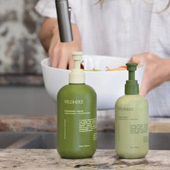 Two Wild Heke skincare bottles on a countertop in a kitchen setting, with a person washing fruit in the background.- Rich gentle lather of Wildheke dishwash liquid cleaning wine glasses next to fresh native green Kawakawa leaves.-THE COSMETIC STORE NZ.