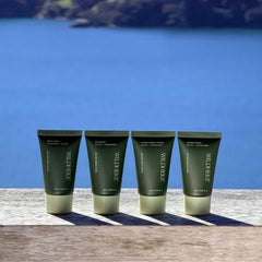 Wildheke natural travel size mini toiletries set 4x25ml sitting neatly inside an unzipped suitcase NZ-Four dark olive green Wild Heart skincare tubes arranged in a row on a wooden surface with a scenic blue ocean and rocky coastline in the background.
