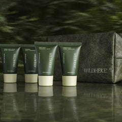 Wildheke Essential Travel Kit 4x25ml mini bottles with pouch at The Cosmetic Store-Four Wildheke body wash tubes in dark green and cream are lined up on a reflective dark surface, with a matching dark green textured pouch behind them against a blurred green background.