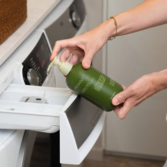 Wildheke Natural Laundry Liquid Horopito and Kawakawa 500ml concentrated pump bottle at The Cosmetic Store NZ.- A person's hands holding an olive green pump bottle of laundry detergent over an open white laundry detergent drawer on a washing machine.