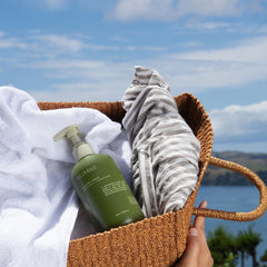 A hand holds a woven brown basket outdoors by the water, containing a white towel, a striped gray-and-white towel, and an olive green pump bottle labeled WILDHEKE.-Gentle plant based laundry liquid cleaning organic cotton towels next to fresh green native Kawakawa leaves.
