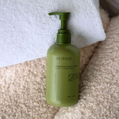 A bottle of Wildheke Magnesium Body Lotion in olive green, placed on a beige textured rug next to a white towel.-Smooth white texture of Wildheke magnesium lotion next to fresh native green Kawakawa leaves-THE COSMETIC STORE NZ.