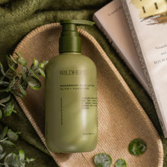 A bottle of WildHERE Magnesium Body Lotion in olive green, styled on a woven tray with green leaves and an open book.-WILDHEKE MAGNESIUM BODY LOTION – Olive + Kawakawa 300ml -The Cosmetic Store NZ.