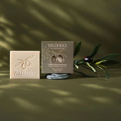 •  Close up texture of Wildheke smooth white squalane solid soap lather next to green botanical herbs
- Wildheke olive & scullion soap shown with a rectangular bar of soap embossed with an olive branch and a matching branded box, styled with olive leaves and a dark olive-brown background with soft shadow overlays.