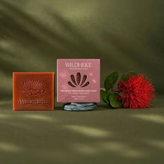 • Close up of rich creamy lather from Wildheke red wine and native Pohutukawa honey soap next to red grapes-THE COSMETIC STORE NZ.- Wildheke Red Wine & Konstuakura Honey Soap shown as a square pink box with the brand name and soap illustration, alongside a brown soap bar embossed with the Wildheke logo and a red flower prop, all arranged on an olive green surface with soft leaf shadows.