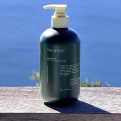 A 500ml pump bottle of Wildheke Shampoo, Olive + Kawakawa, placed on a weathered wooden ledge with a bright blue ocean background.-  Eco friendly Wildheke scalp care shampoo 500ml sitting on a marble shower ledge NZ