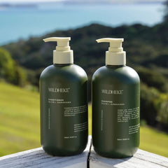 Two dark green pump bottles of WILDHEKE hair care products, one labeled conditioner and one labeled shampoo, standing on a light-colored wooden surface with a blurred outdoor background of green hills and blue water.- Luxurious sulfate free lather of Wildheke shampoo next to fresh green native Kawakawa leaves-THE COSMETIC STORE NZ.
