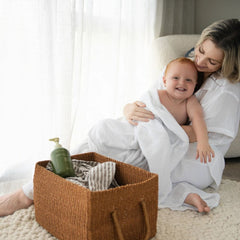 A woman in a white robe holds a smiling baby wrapped in a white towel on a light rug, with a woven brown basket and a green pump bottle nearby in a bright indoor setting.- Wildheke Natural Laundry Liquid Horopito and Kawakawa 500ml concentrated pump bottle at The Cosmetic Store NZ.