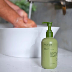 Wildheke Natural Hand Wash Kawakawa and Kanuka 300ml pump bottle at The Cosmetic Store NZ-A bottle of WILDFEKE hand wash in a light olive green color with a matching green pump dispenser, placed on a white marble countertop in front of a blurred background showing a hand washing in a sink.