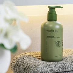 • Clear gentle lather of Wildheke hand wash next to green native Kawakawa leaves and Kanuka twigs
-THE COSMETIC STORE NZ.- A green pump bottle of WILDHEKE hand wash labeled 'KAWAKAWA + KANUKA' resting on a textured gray towel with a soft-focus background.