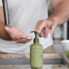 Wildheke hydrating natural hand wash 300ml sitting next to a modern matte black sink NZ-THE COSMETIC STORE NZ.-A person in a white tank top pumps a bottle of Wild Haze Hand Wash onto their hand at a kitchen sink.