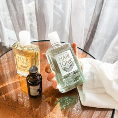 Two bottles of hair tonic are shown on a reflective copper-toned table with a hand holding one clear bottle and another bottle standing beside it. A white towel is also visible, and soft white curtains hang in the background.- Yanagiya Hair Tonic Citrus 240ml- THE COSMETIC STORE NZ